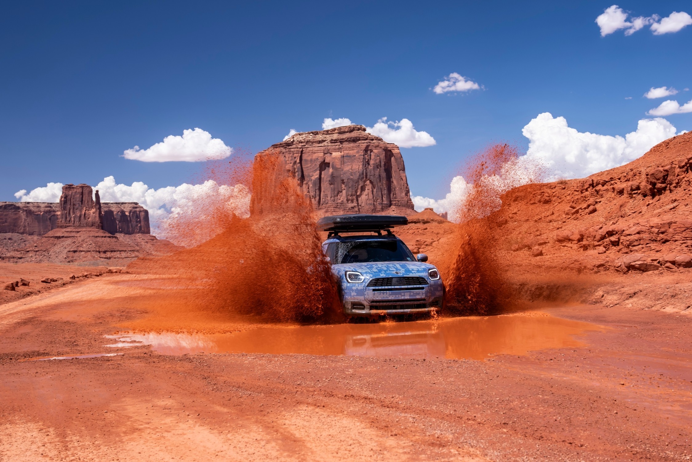 MINI Countryman a atravessar uma poça de lama num terreno desértico, com salpicos  de água e formações rochosas ao fundo 